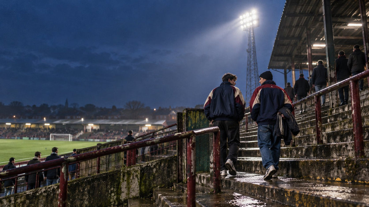 Two friends in retro track jackets climbing wet terrace steps at a floodlit football ground at dusk