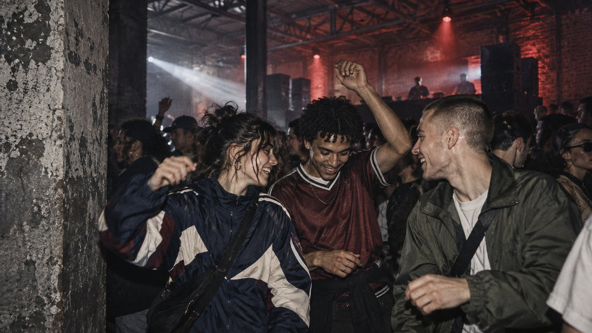 Three friends laughing and dancing in a packed warehouse rave, DJ and red lights behind